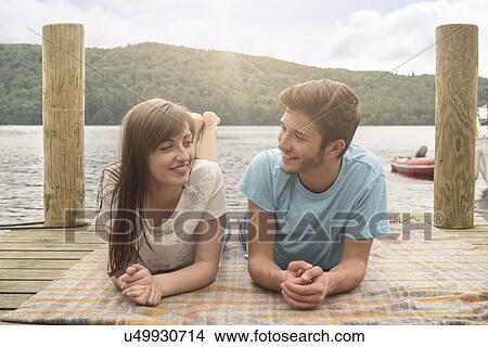 Picture - Young smiling couple lying on blanket on lakeside jetty. Fotosearch