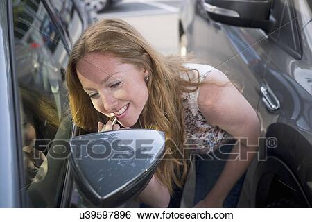 Stock Photograph - Young woman applying lipstick whilst looking in car wind mirror. Fotosearch