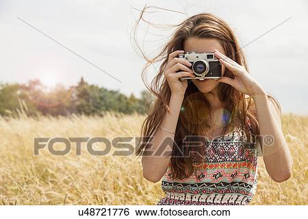 Stock Photograph - Young woman taking photo with vintage camera in field. Fotosearch