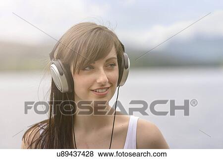 Stock Photo - Young woman wearing headphones at lakeside under bright sunlight. Fotosearch