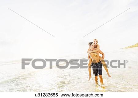 Stock Photography - Boyfriend walking in surf carrying girlfriend piggyback. Fotosearch