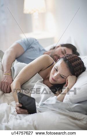 Stock Photo - Couple in bed, woman reading on digital tablet and man sleeping. Fotosearch
