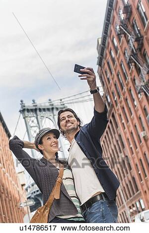 Stock Photo - Couple taking selfie on street, Brooklyn Bridge in background. Fotosearch