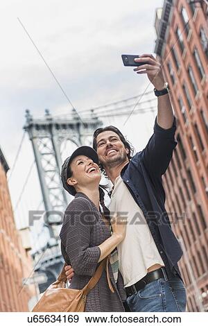Stock Photo - Couple taking selfie on street, Brooklyn Bridge in background. Fotosearch