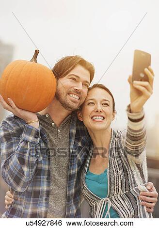 Stock Photograph - Couple taking selfie with mobile phone, man holding pumpkin. Fotosearch