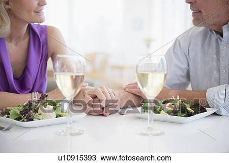 Stock Image - Cropped view of couple holding hands in restaurant with glasses of white wine in foreground. Fotosearch