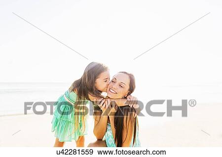 Stock Photo - Girl (6-7) kissing her mother on cheek on beach. Fotosearch