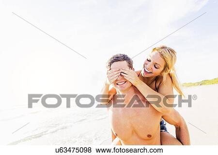 Stock Photo - Girlfriend riding piggyback on beach, closing boyfriend's eyes with palms. Fotosearch
