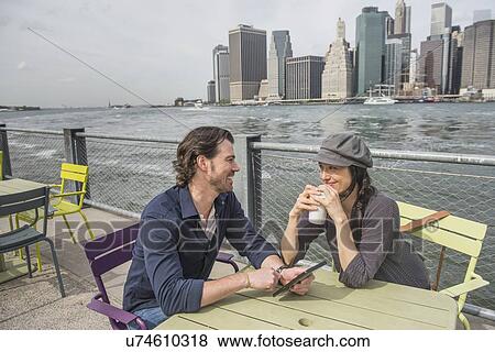 Stock Photo - Happy couple sitting and using tablet pc and drinking coffee with cityscape in background. Fotosearch