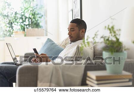 Picture - Man sitting in living room, using laptop and cell phone. Fotosearch