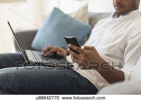 Stock Image - Man sitting in living room, using laptop and cell phone. Fotosearch