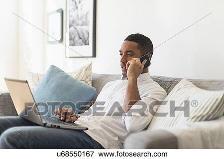 Man sitting in living room, using laptop and cell phone View Large Photo Image Stock Photo - Man sitting in living room, using laptop and cell phone. Fotosearch