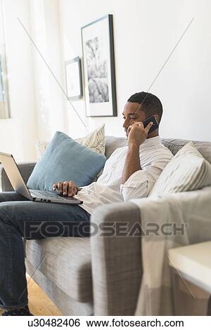 Stock Photograph - Man sitting in living room, using laptop and cell phone. Fotosearch
