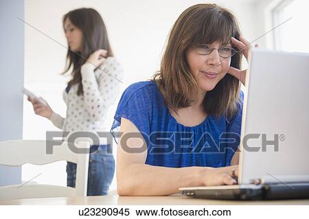 Stock Photography - Mother using laptop, daughter (14-15) using mobile phone in background. Fotosearch
