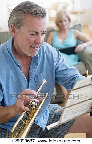 Stock Photo - Portrait of senior man holding trumpet looking at music stand with woman on sofa in background. Fotosearch