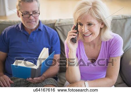 Stock Photograph - Portrait of woman talking on phone and man reading book, sitting on sofa. Fotosearch