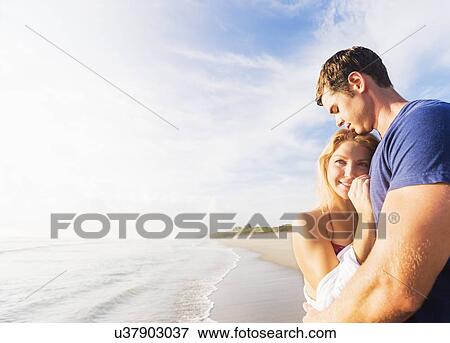 Stock Photo - Portrait of young couple embracing on sandy beach, against background of coastline. Fotosearch