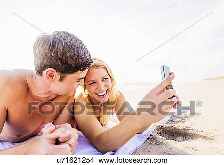 Picture - Portrait of young couple lying on blanket on beach, using smart phone. Fotosearch