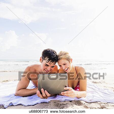 Stock Image - Portrait of young couple lying on blanket on beach side by side, using digital tablet. Fotosearch