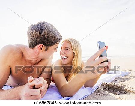 Stock Photography - Portrait of young couple lying on blanket on beach, using smart phone. Fotosearch