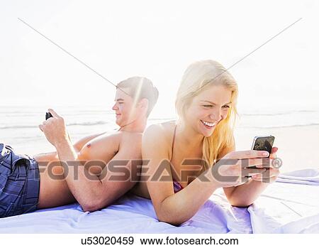 Stock Photo - Portrait of young couple relaxing on beach, using smart phones. Fotosearch