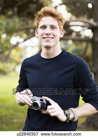 Portrait of young man holding camera in park View Large Photo Image Stock Photo - Portrait of young man holding camera in park. Fotosearch