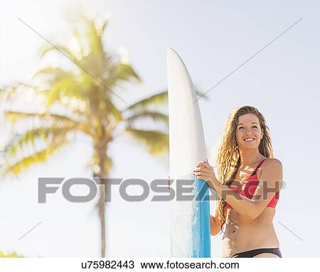 Stock Image - Portrait of young woman holding surfboard on beach. Fotosearch