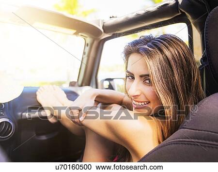 Stock Image - Portrait of young woman sitting in car with legs on dashboard. Fotosearch