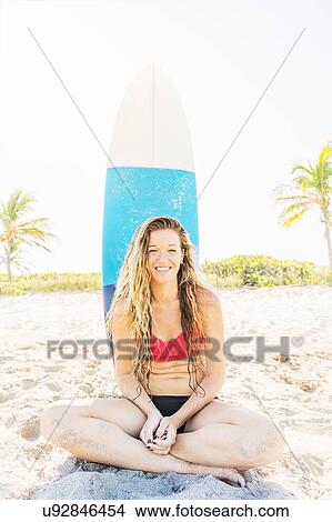 Portrait of young woman sitting on beach with surfboard in background View Large Photo Image Picture - Portrait of young woman sitting on beach with surfboard in background. Fotosearch