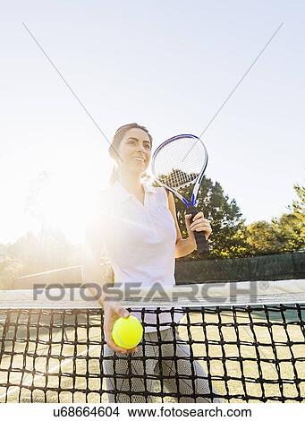 Portrait of young woman standing by net, holding tennis ball and tennis racket View Large Photo Image Picture - Portrait of young woman standing by net, holding tennis ball and tennis racket. Fotosearch