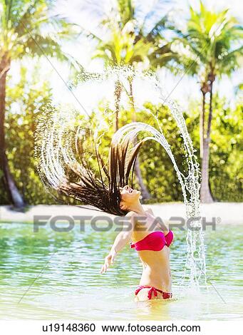 Stock Image - Portrait of young woman wearing bikini tossing hair and splashing water in tropical lagoon. Fotosearch