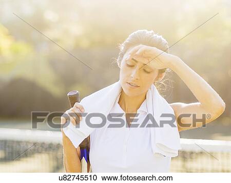 Stock Image - Portrait of young woman with towel and tennis racket wiping forehead with back of hand. Fotosearch