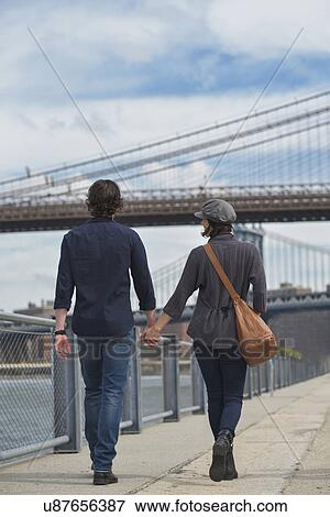 Rear view of couple walking on promenade, Brooklyn Bridge in background View Large Photo Image Stock Photo - Rear view of couple walking on promenade, Brooklyn Bridge in background. Fotosearch