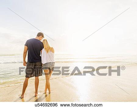 Stock Photograph - Rear view of young couple standing side by side on sandy beach, looking at sea. Fotosearch