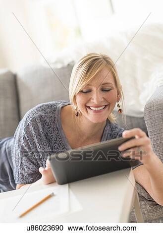 Stock Image - Smiling woman lying down on sofa with tablet pc. Fotosearch
