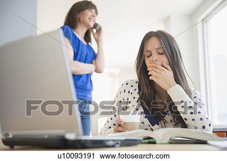 Stock Image - Tired teenage girl (14-15) yawning in front of textbook, mother on phone in background. Fotosearch