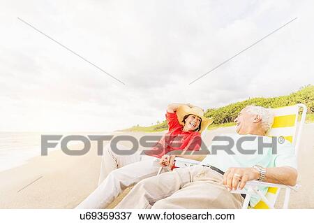 Stock Photo - View of couple sitting in lounge chairs on beach and laughing. Fotosearch