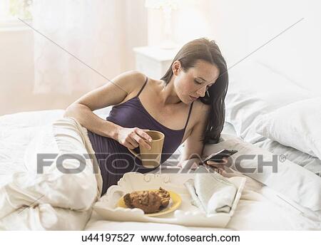 Stock Photo - Woman having breakfast in bed, using mobile phone. Fotosearch