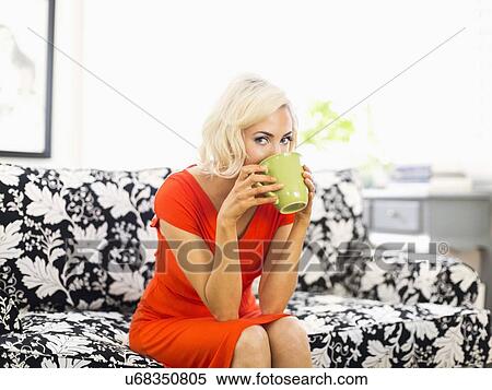 Stock Photography - Woman in red dress sitting on sofa and drinking coffee. Fotosearch