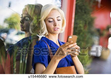 Stock Image - Woman leaning against window and using phone. Fotosearch