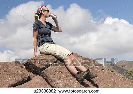 Stock Image - Woman sitting on rock and drinking from bottle. Fotosearch