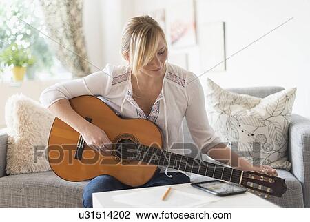Stock Image - Woman sitting on sofa and playing acoustic guitar. Fotosearch