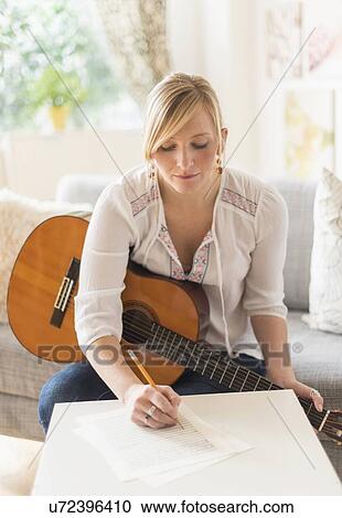 Stock Image - Woman sitting on sofa with acoustic guitar and writing. Fotosearch