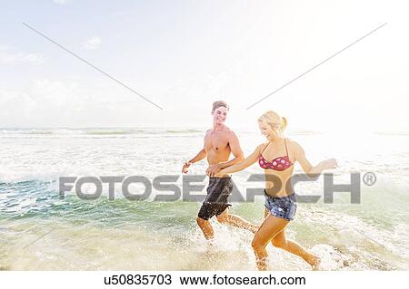 Stock Image - Young couple running in surf holding hands. Fotosearch