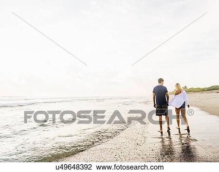 Stock Image - Young couple walking along surf line of sandy beach, holding hands. Fotosearch