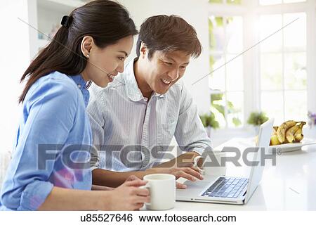 Stock Photograph - Asian Couple Looking at Laptop In Kitchen. Fotosearch