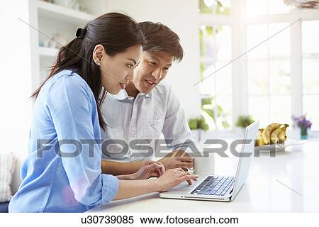Stock Photography - Asian Couple Looking at Laptop In Kitchen. Fotosearch