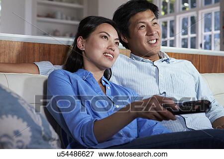 Stock Photo - Asian Couple Sitting On Sofa Watching TV Together. Fotosearch
