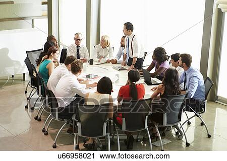 Stock Image - Businessman Addressing Meeting Around Boardroom Table. Fotosearch