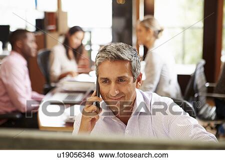 Businessman On Phone At Desk With Meeting In Background View Large Photo Image Stock Photo - Businessman On Phone At Desk With Meeting In Background. Fotosearch